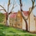 A painting of a traditional stone building with multiple windows and a red roof. In the foreground, there are large trees with bare branches and some green foliage. The grass is lush and green, leading up to the building.