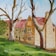 A painting of a traditional stone building with multiple windows and a red roof. In the foreground, there are large trees with bare branches and some green foliage. The grass is lush and green, leading up to the building.