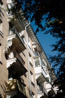 A multi-story residential building with brickwork and balconies. Satellite dishes and air conditioning units can be seen on the exterior, with overhanging structures above each balcony. The scene is framed by tree branches with leaves, against a backdrop of a clear blue sky.