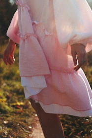 A model wearing a flowing pastel pink dress adorned with small heart motifs, standing in a sunlit garden.