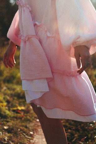 A flowing pastel pink dress with a cinched waist, worn by a woman walking through a sunlit garden pathway.