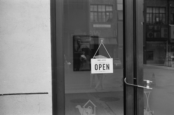 A black and white photograph of a glass door with a sign that reads 'WE ARE DEFINITELY OPEN, COME ON IN.' The door handle is visible on the right side, and reflections of buildings and street scenes can be seen in the glass.