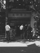 A group of people, including a child sitter, standing or sitting on stools in front of a building entrance with Chinese characters on the signage. The scene is captured in black and white, and there are shadows of trees visible on the ground and building.