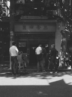 A group of people, including a child sitter, standing or sitting on stools in front of a building entrance with Chinese characters on the signage. The scene is captured in black and white, and there are shadows of trees visible on the ground and building.