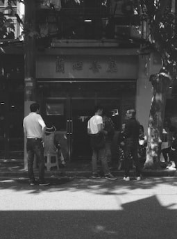 A group of people, including a child sitter, standing or sitting on stools in front of a building entrance with Chinese characters on the signage. The scene is captured in black and white, and there are shadows of trees visible on the ground and building.