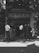A group of people, including a child sitter, standing or sitting on stools in front of a building entrance with Chinese characters on the signage. The scene is captured in black and white, and there are shadows of trees visible on the ground and building.
