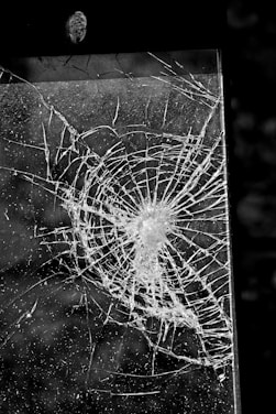 A cracked or shattered glass panel with a complex pattern of fractures radiating from a central point. The image is in black and white, highlighting the intricate lines and textures created by the breakage.