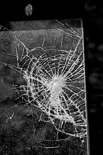 A cracked or shattered glass panel with a complex pattern of fractures radiating from a central point. The image is in black and white, highlighting the intricate lines and textures created by the breakage.