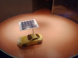 A small yellow toy car is equipped with a solar panel mounted on a pole, set on a circular surface with an orangish hue. The setting is enclosed and illuminates the solar panel prominently.