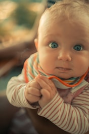 A baby with large, curious blue eyes is looking directly at the camera. The child has blonde hair and is wearing a striped shirt and a colorful bib. The background is softly blurred, creating a warm and intimate atmosphere.
