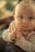 A baby with large, curious blue eyes is looking directly at the camera. The child has blonde hair and is wearing a striped shirt and a colorful bib. The background is softly blurred, creating a warm and intimate atmosphere.