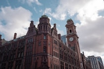 A historic brick building with intricate architectural details, featuring a prominent clock tower labeled 'Kimpton'. The sky is partly cloudy and the scene is urban, with modern buildings visible in the background.