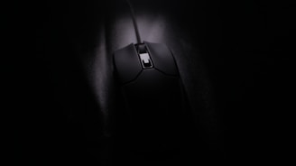 A black computer mouse with a cord, positioned on a dark surface with low lighting. The focus is on the details of the mouse, highlighting its smooth texture and sleek design.
