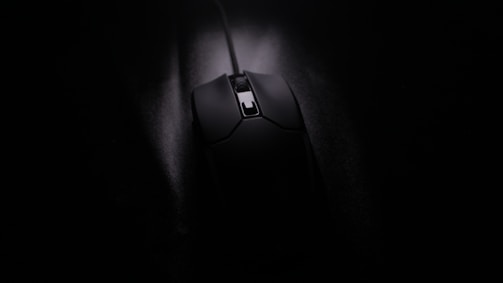 A black computer mouse with a cord, positioned on a dark surface with low lighting. The focus is on the details of the mouse, highlighting its smooth texture and sleek design.
