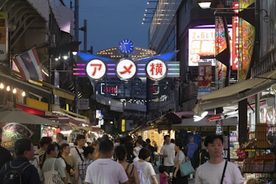 A bustling street market scene under evening light with illuminated signs. People are walking and engaging with various stalls, which display colorful banners and products. A large, lit-up sign with Japanese characters hangs prominently in the center.