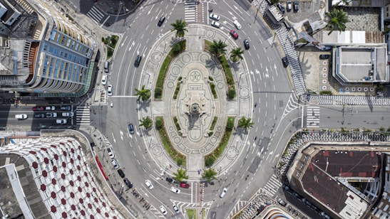 Aerial view of a large roundabout with a monument in the center. The roundabout has well-defined lanes with cars navigating around it. Surrounding the roundabout are various buildings, including a tall, multi-storeyed building with colorful windows and a building with a geometric white and red roof pattern. There are trees and green patches within the roundabout and along the outer area.