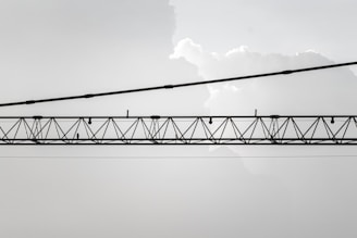 An industrial crane with a lattice structure is suspended horizontally against a backdrop of light, cloudy sky. The crane is composed of metal beams and cables, and it stretches across the image from left to right.
