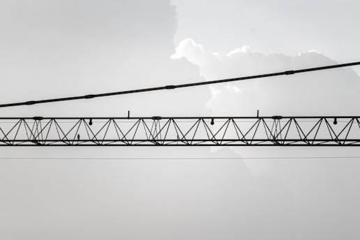 An industrial crane with a lattice structure is suspended horizontally against a backdrop of light, cloudy sky. The crane is composed of metal beams and cables, and it stretches across the image from left to right.