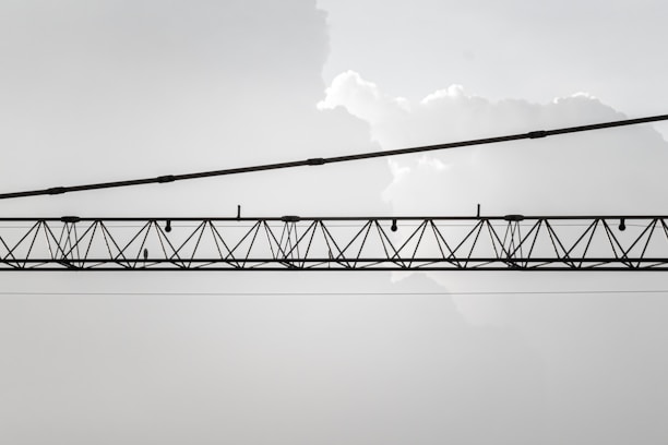 An industrial crane with a lattice structure is suspended horizontally against a backdrop of light, cloudy sky. The crane is composed of metal beams and cables, and it stretches across the image from left to right.