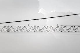An industrial crane with a lattice structure is suspended horizontally against a backdrop of light, cloudy sky. The crane is composed of metal beams and cables, and it stretches across the image from left to right.