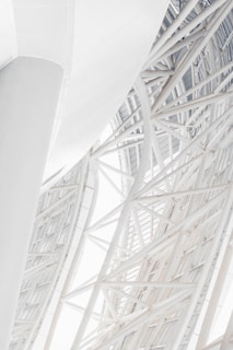 A close-up view of a modern architectural structure featuring intersecting steel beams and geometric patterns. The structure appears to be part of a large building, with a curved design and a clean, white aesthetic. The beams create an intricate lattice, giving the structure a complex and artistic look.