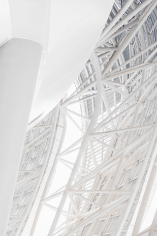 A close-up view of a modern architectural structure featuring intersecting steel beams and geometric patterns. The structure appears to be part of a large building, with a curved design and a clean, white aesthetic. The beams create an intricate lattice, giving the structure a complex and artistic look.