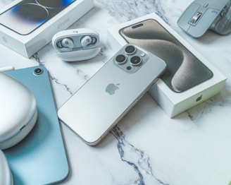 An arrangement of modern technology items including a smartphone, laptop, earbuds, and a mouse. The smartphone is prominently displayed with its box, featuring a sleek design and multiple cameras. The earbuds are neatly placed in an open case. A laptop and a wireless mouse are also visible, all set against a marble surface.