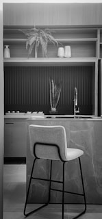 A modern kitchen interior with a marble countertop and a high chair positioned at the island. Above the counter is a sink with a sleek, metallic faucet. The background features open shelving with decorative items including a potted fern and vases. The space is lit naturally, highlighting the textured surfaces and clean lines.