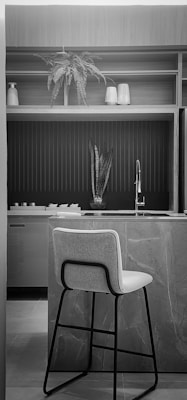 A modern kitchen interior with a marble countertop and a high chair positioned at the island. Above the counter is a sink with a sleek, metallic faucet. The background features open shelving with decorative items including a potted fern and vases. The space is lit naturally, highlighting the textured surfaces and clean lines.
