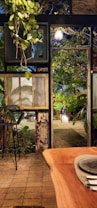 An outdoor seating area featuring rustic wooden architecture and abundant greenery is illuminated by a hanging light. The scene includes a wooden table with plates and cutlery, surrounded by tall plants and antique-style window frames. The ambiance suggests a cozy, nature-infused dining experience.