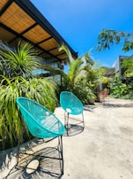 Two vibrant turquoise chairs with a woven design are placed on a concrete patio surrounded by lush green tropical plants. A wooden pergola and modern building with large windows and a bamboo-shaded roof are visible in the background under a clear blue sky.