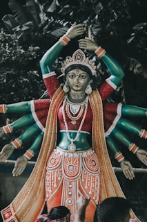 A sculpture of a deity with multiple arms, adorned with intricate jewelry and garments, is placed against a backdrop of lush greenery. The statue features a rich palette of colors and detailed craftsmanship.