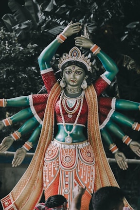 A sculpture of a deity with multiple arms, adorned with intricate jewelry and garments, is placed against a backdrop of lush greenery. The statue features a rich palette of colors and detailed craftsmanship.