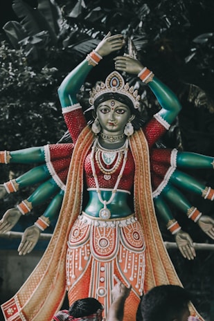 A sculpture of a deity with multiple arms, adorned with intricate jewelry and garments, is placed against a backdrop of lush greenery. The statue features a rich palette of colors and detailed craftsmanship.