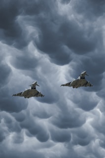Two fighter jets fly through a dramatic, cloudy sky with swirling, dark gray clouds creating a turbulent background.