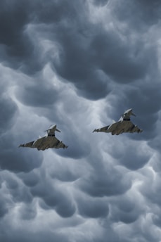 Two fighter jets fly through a dramatic, cloudy sky with swirling, dark gray clouds creating a turbulent background.