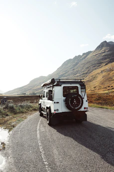 An old Bronco perched on a narrow mountain trail surrounded by rugged terrain and pine trees under a clear blue sky.