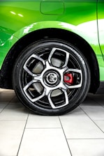 A detailed close-up view of a car wheel with a black alloy rim and a prominent red brake caliper. The car body is a vibrant green, and there is a reflection on its surface. The tire has the brand name 'Continental' visible. The background consists of tiled flooring in a light color.