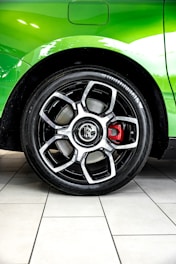 A detailed close-up view of a car wheel with a black alloy rim and a prominent red brake caliper. The car body is a vibrant green, and there is a reflection on its surface. The tire has the brand name 'Continental' visible. The background consists of tiled flooring in a light color.