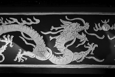A detailed depiction of a dragon is shown in relief against a dark background. The dragon has a long, winding body and is adorned with intricate patterns, including scales and stylized flames. The scene includes elements resembling wisps of clouds and a circular object, possibly representing the sun.