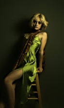 A person with blonde hair is leaning against a wooden stool, wearing a green satin dress with a thigh-high slit. A brown shawl drapes over one shoulder, creating a sophisticated yet relaxed look. The dark, muted background adds to the overall mood.