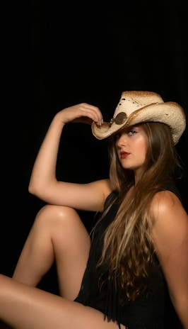 A woman with long hair is sitting in a relaxed pose while wearing a cowboy hat. The background is dark, highlighting her features and clothing.