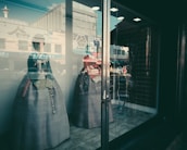 Two mannequins are displayed in a store window wearing traditional Korean hanboks. The reflection of street activity, including people and vehicles, is visible on the glass, adding an urban backdrop. The interior of the shop is dimly lit, contrasting with the bright scene outside.