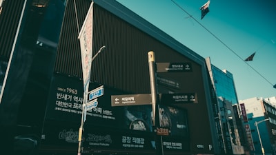 A street scene in an urban area featuring a modern building with large signage in Korean. Various signposts are visible pointing towards local attractions like Jagalchi Station and Jagalchi Market. The building's facade is dominated by vertical lines and Korean text along with English translations for directions.