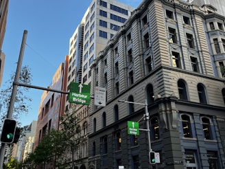 A city street with tall classic and modern buildings, traffic lights, and signs directing towards the Harbour Bridge. Trees are visible along the street, and the sky is clear and blue.