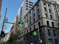 A city street with tall classic and modern buildings, traffic lights, and signs directing towards the Harbour Bridge. Trees are visible along the street, and the sky is clear and blue.