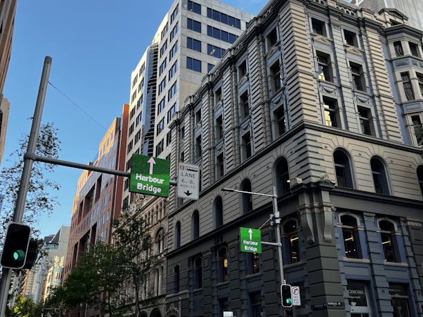 A city street with tall classic and modern buildings, traffic lights, and signs directing towards the Harbour Bridge. Trees are visible along the street, and the sky is clear and blue.