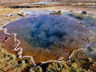 A geothermal hot spring with a vivid blue pool at the center, surrounded by earthy tones and patches of green vegetation. The edges of the pool have mineral deposits creating intricate patterns. In the background, a barren landscape stretches out with people visible in the distance, indicating a natural tourist site.