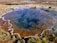 A geothermal hot spring with a vivid blue pool at the center, surrounded by earthy tones and patches of green vegetation. The edges of the pool have mineral deposits creating intricate patterns. In the background, a barren landscape stretches out with people visible in the distance, indicating a natural tourist site.