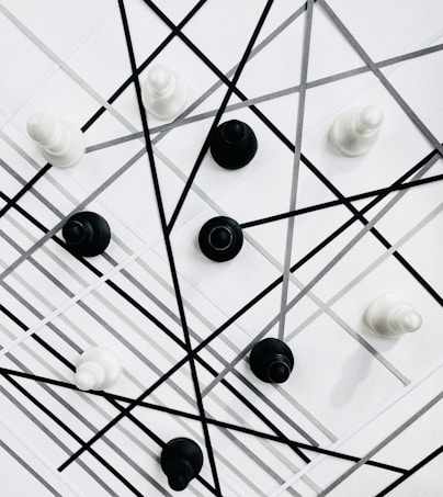 Chess pieces are arranged on a geometric background with intersecting black and gray lines. The pieces are predominantly black and white, creating a contrast against the patterned backdrop.